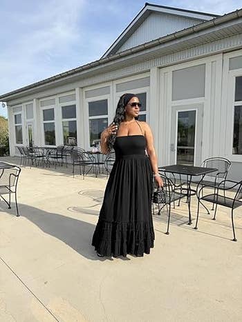 Person wearing a flowing black dress stands outdoors, near a white building with glass tables and chairs, posing confidently