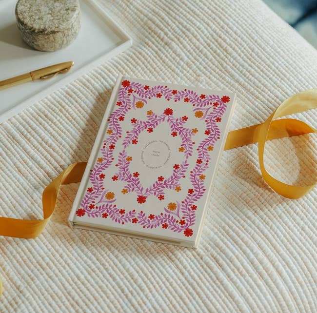 A decorative notebook with a floral screen plan and a yellowish ribbon bookmark is placed connected a textured surface, alongside a pen and a stone-paperweight