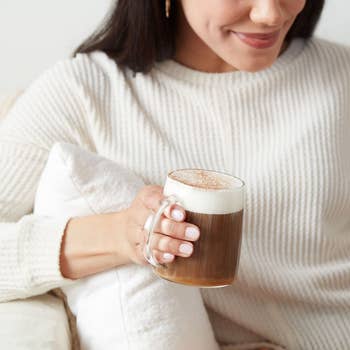 Woman in white sweater holds a frothy coffee mug while seated on a couch, smiling slightly