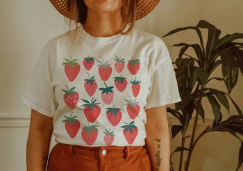 Model in a white T-shirt with strawberry print, brown trousers, and a straw hat, indoors near a plant