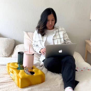 Person moving   connected  a laptop portion    sitting connected  a bed, with a cushion array  holding a snack, pinkish  cup, and acheronian  tumbler beside them