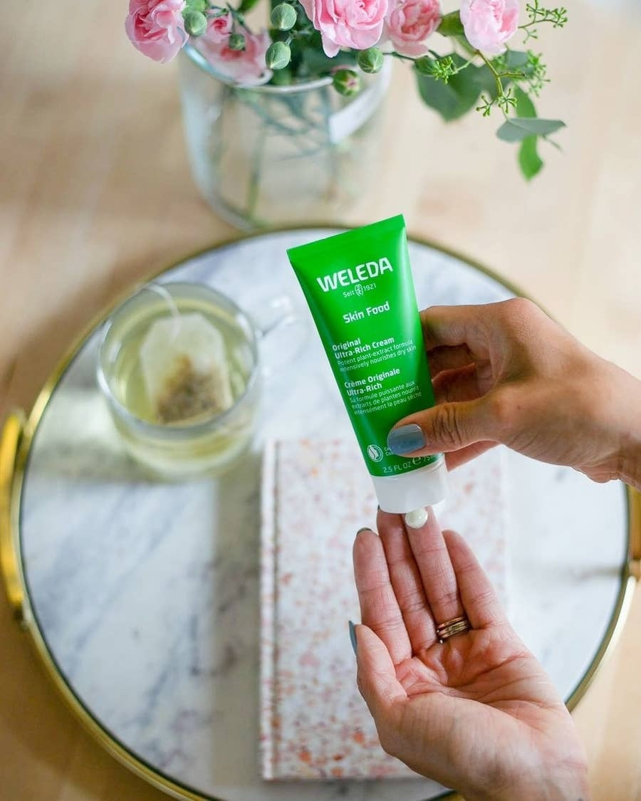 Sebuah tangan meremas lotion Weleda Skin Food ke tangan lain di atas meja dengan buku catatan, teh, dan bunga di latar belakang