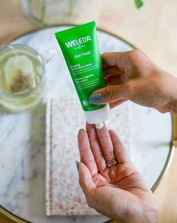 Sebuah tangan meremas lotion Weleda Skin Food ke tangan lain di atas meja dengan buku catatan, teh, dan bunga di latar belakang