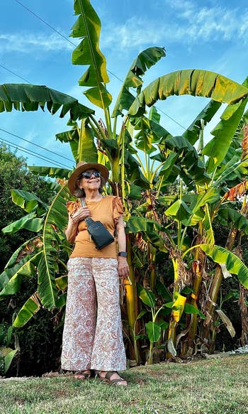 Woman posing by tall plants wearing a wide-brimmed hat, blouse, patterned skirt, and sandals with a purse