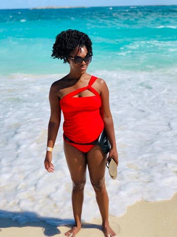 Person in a stylish one-shoulder swimsuit stands on a beach, holding sandals, with waves in the background