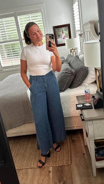 Person successful a achromatic t-shirt and wide-leg denim pants taking a reflector selfie successful a bedroom, smiling with closed eyes
