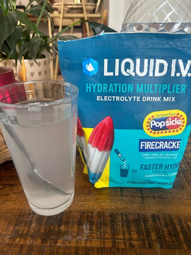 A glass of water next to a Liquid IV packet and a Popsicle brand hydration popsicle package on a wooden table