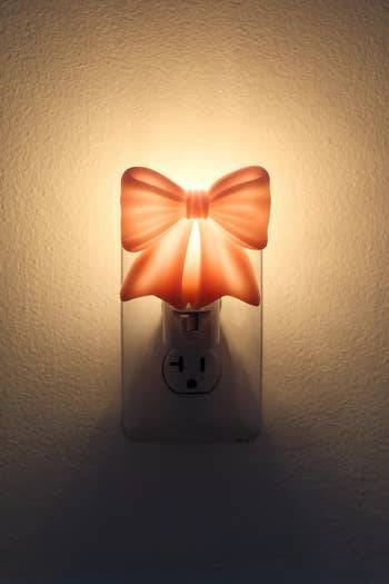 Nightlight shaped like a bow, plugged into a wall outlet, softly illuminating the surrounding area