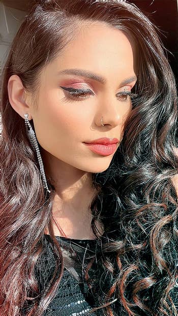 Person with wavy hair and hoop earrings wearing a black outfit, showcasing a makeup look with highlighted eyes and red lipstick