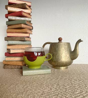 mug of tea on the green wooden book coaster next to a teapot and a giant stack of the coasters