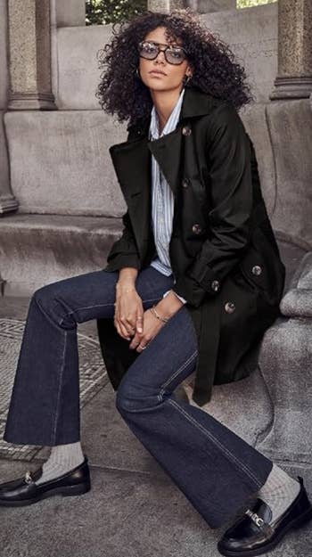 Person seated outdoors wearing a stylish trench coat, flared jeans, and loafers, with sunglasses and curly hair, appearing relaxed and fashionable