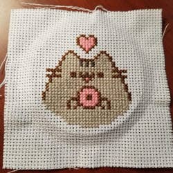 reviewer's cross stitch of pusheen's upper body holding a donut with a heart over her head