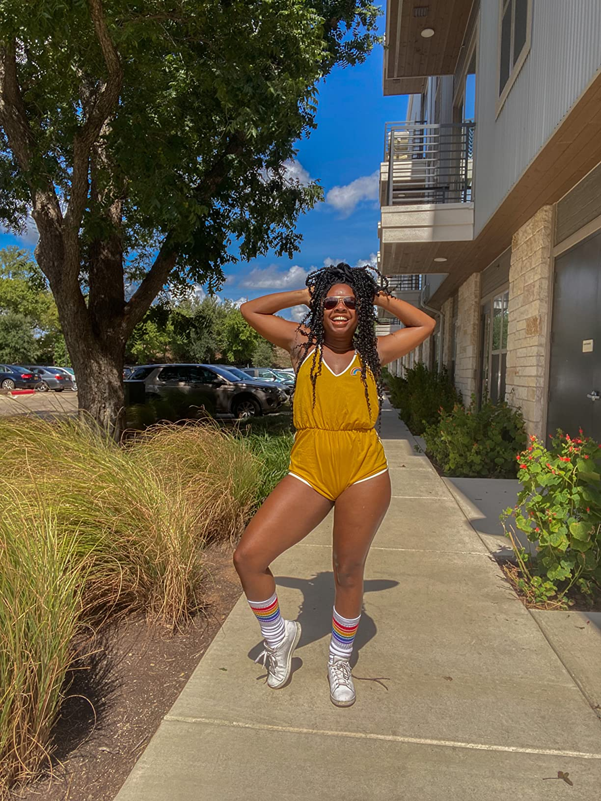 Person posing outdoors successful  a stylish romper and sneakers, wearing sunglasses and socks with stripes, lasting  connected  a paved sidewalk beside writer  and trees