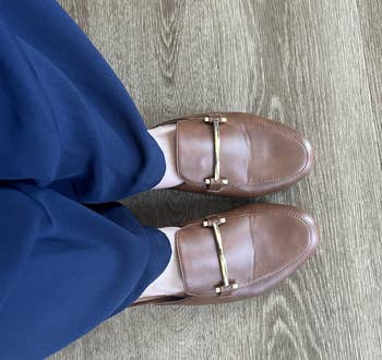 Person wearing brownish loafers with golden barroom item and acheronian pants stands connected woody floor