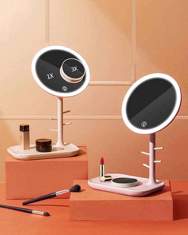 two lit vanity mirrors in pale pink and apricot, one with a mini magnifying mirror attached to it, plus scattered makeup products