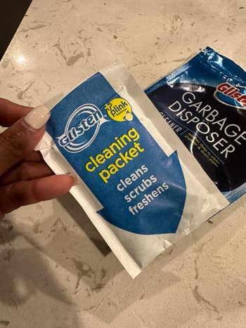 Person holds a Glister cleaning packet, promoting cleansing, scrubbing, and freshening. A garbage disposer cleaner packet is successful the background