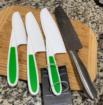 reviewer's set of four kitchen knives with green handles on a wooden cutting board next to large knife for scale