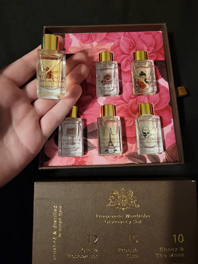 Set of six tiny  fragrance bottles with floral packaging, manus  holding 1  bottle. Displayed connected  a container  labeled 