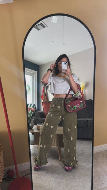 Person taking a reflector  selfie, wearing a cropped top, floral pants with ruffles, and holding a colorful bag