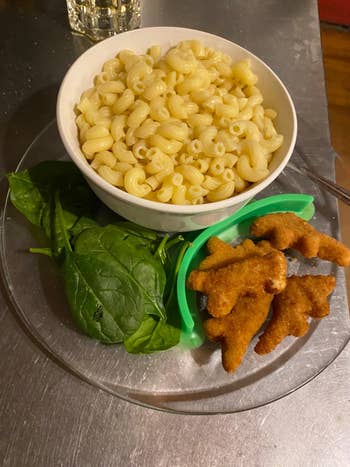 Bowl of macaroni with spinach leaves and dinosaur-shaped chickenhearted  nuggets connected  a plate
