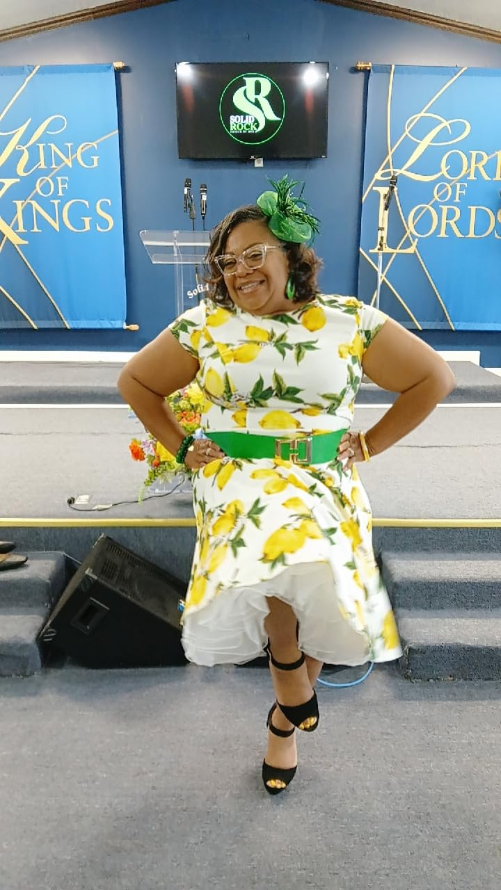 Person wearing a lemon-print dress with a green belt and fascinator, smiling and posing indoors on a stage