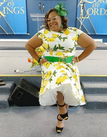 Person wearing a lemon-print dress with a green belt and fascinator, smiling and posing indoors on a stage