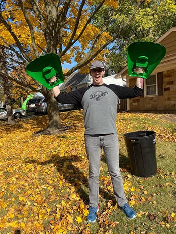 Person wearing a casual shirt and jeans, holding oversized rake hands in a yard covered with leaves