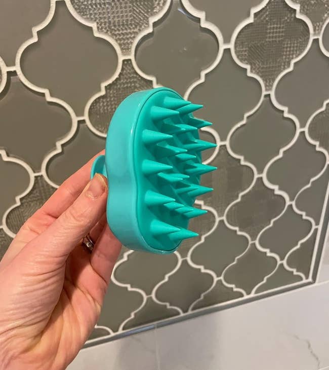 Hand holding a turquoise scalp massaging brush against a patterned tile background