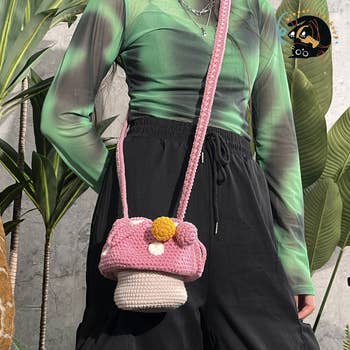 Model wearing a green top, black pants, and holding a pink crochet handbag