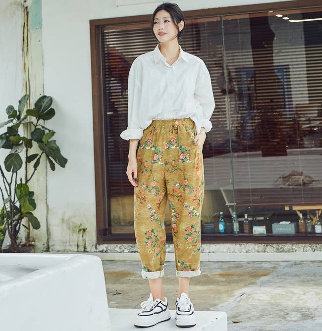 Person in casual attire with a white shirt and floral pants, standing outdoors in front of a shop window