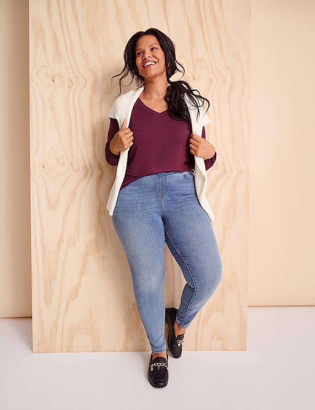 Woman successful casual outfit, wearing a maroon top, achromatic sweater vest, jeans, and loafers, leaning against a woody wall, smiling