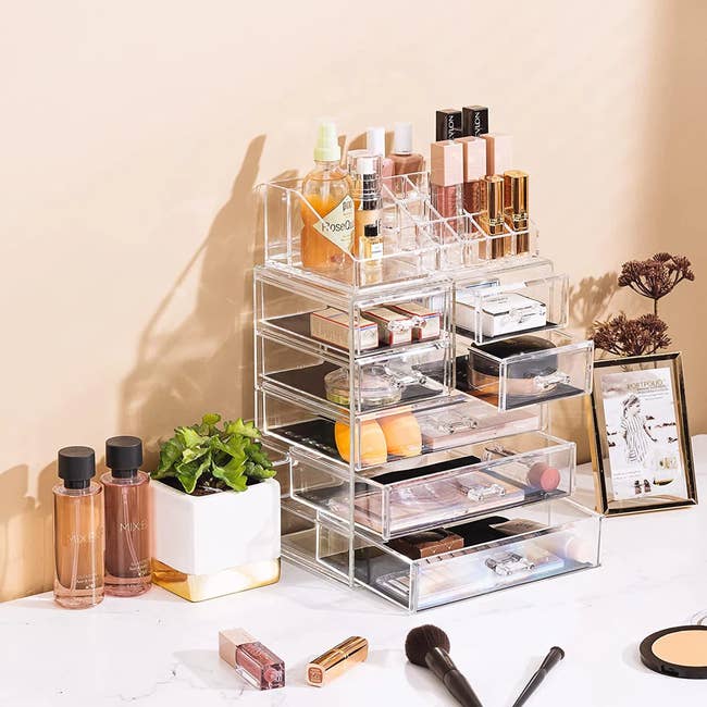 Clear acrylic makeup organizer with drawers, containing lipsticks, perfumes, and skincare products, placed on a vanity alongside brushes and plants