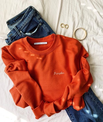 the orange sweatshirt on top of a paid of jeans, with some jewelry nearby