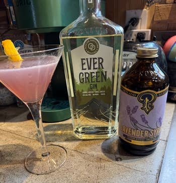 Cocktail adjacent  to a vessel  of Ever Green Gin and lavender syrup connected  a countertop, with a citrus  twist garnish