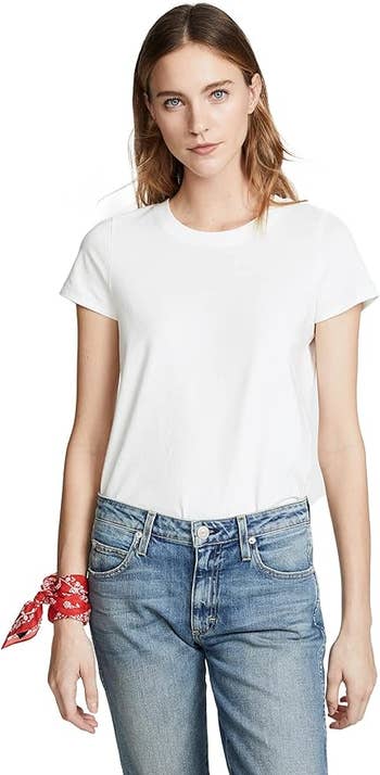 Model in a plain white tee and blue jeans with a red accessory on wrist