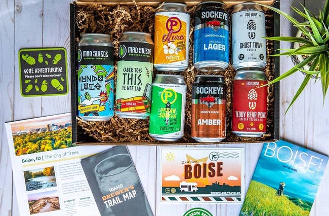 A variety of craft beer cans in a gift box with Idaho-themed items, including maps and a postcard, are arranged on a wooden surface