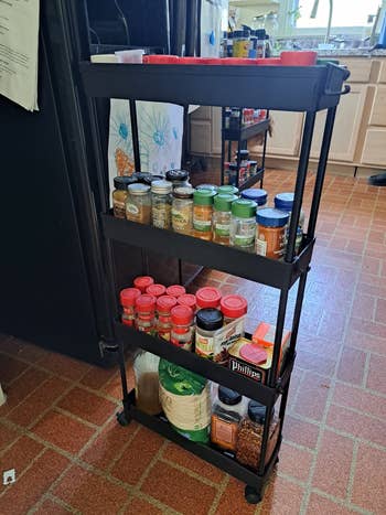 Rolling kitchen cart with three shelves, holding various spices, storage jars, and kitchen items in a tiled kitchen setting