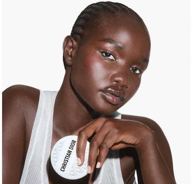 Model poses with Dior product, holding it beside their face, wearing a sleeveless top