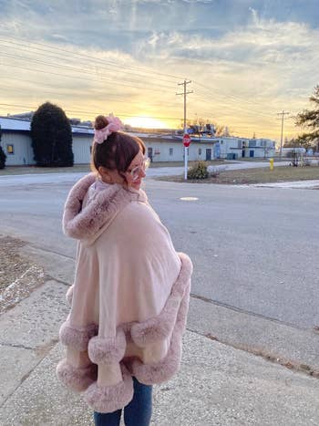 Person outdoors wearing a cozy cape with fur trim, glasses, and a bun, against an concern   backdrop astatine  sunset. Fashionable and casual thoroughfare  style