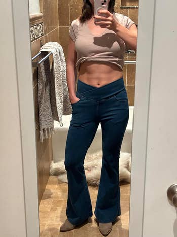 A reviewer in a bathroom mirror selfie wearing a short-sleeve crop top and high-waisted flare jeans with a V-cross waistband, showing off their midriff