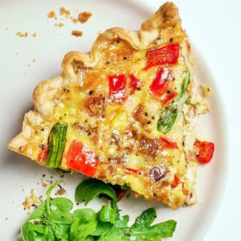 Slice of vegetable quiche with red peppers and greens on a plate, accompanied by a side of fresh salad greens