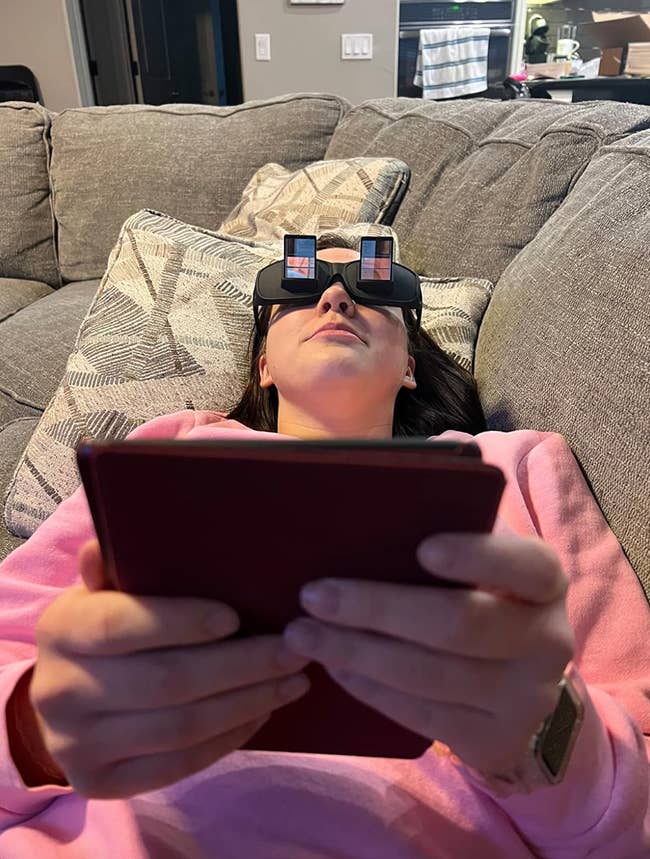 Reviewer laying horizontally on a couch while wearing glasses that let them read a propped up book 