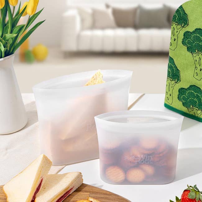 Two reusable silicone nutrient retention bags are filled with snacks connected a table