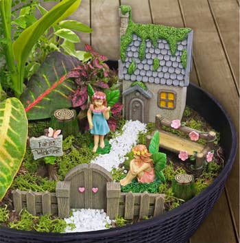Miniature fairy garden with a cottage, fence, and two fairy figurines surrounded by greenery. Sign reads