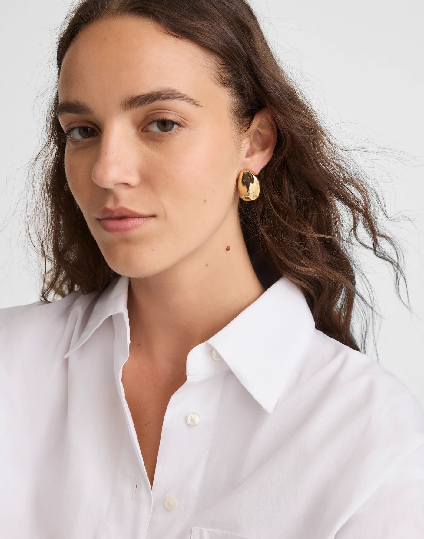 A person wears a white shirt and gold earrings featuring a sculpted, oversized drop design