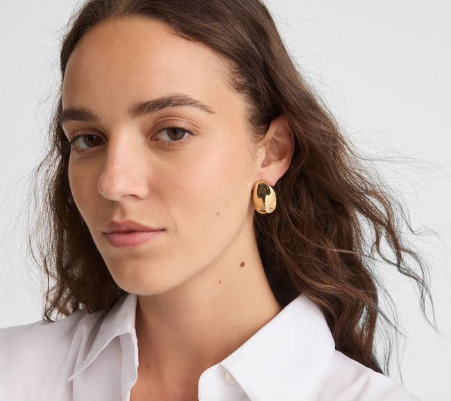 A person wears a white shirt and gold earrings featuring a sculpted, oversized drop design