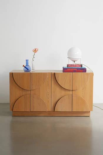 Modern wooden sideboard with geometric design, topped with a round lamp, books, and a blue vase holding a single flower