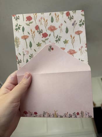 A manus  holds a floral-patterned greeting paper  with a partially inserted airy   envelope, showcasing a nature-themed design