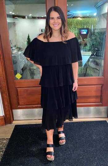 reviewer in a tiered off-the-shoulder black dress and open-toe sandals standing indoors