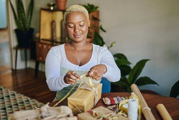A person with short hair wraps a gift at a table, surrounded by wrapping supplies and plants in the background